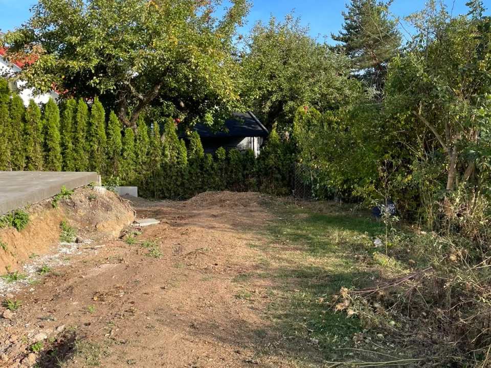 Weitgehend geräumte und abgetragene Gartenfläche neben einer erhöhten Betonplatte mit Hecken und Bäumen im Hintergrund