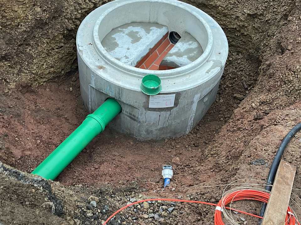 Runder Betonschacht mit angeschlossener grüner Rohrleitung in einer tief ausgehobenen Baugrube