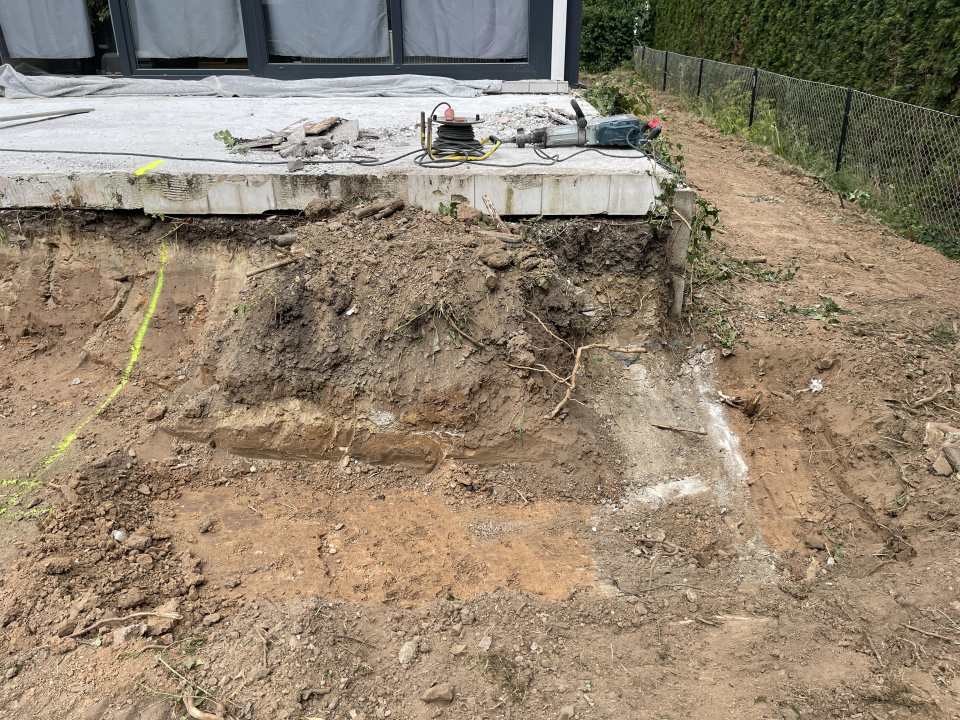 Ausgehobener Hangbereich unter einer bestehenden Betonterrasse mit freigelegtem Erdreich und Wurzeln