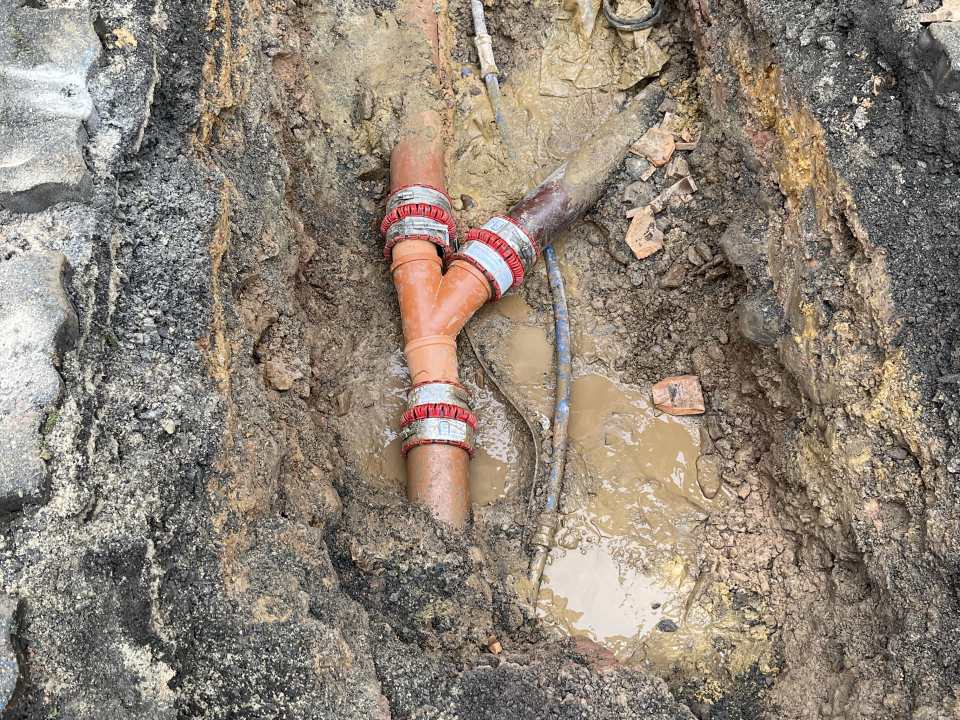 Freigelegter Rohrleitungsanschluss mit orangefarbenen Formteilen und Kupplungen in einem Erdgraben