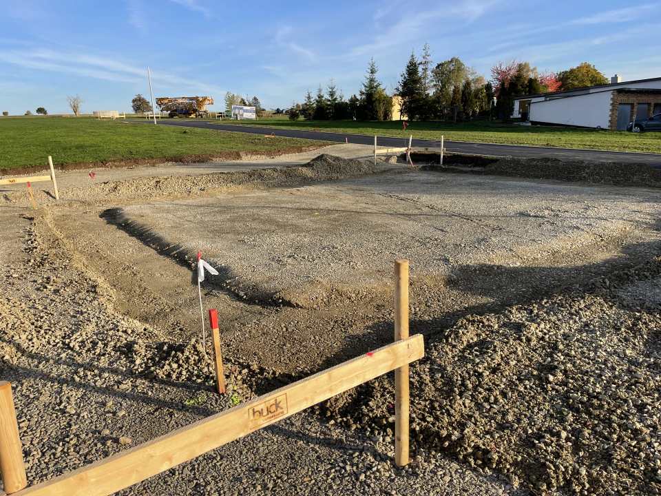 Große vorbereitete Schotterfläche mit Holzabsteckungen und Höhenmarkierungen neben einer neuen Asphaltstraße