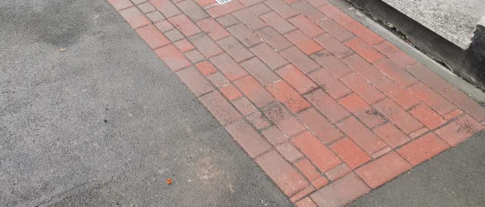 Rechteckige rote Pflasterfläche mit integriertem Metallrost vor einem Gebäudeeingang neben Asphalt