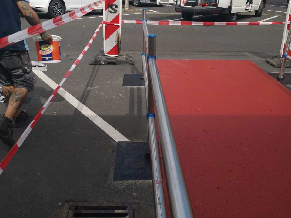 Abgesperrte Zugangsrampe mit roter Beschichtung, Edelstahlgeländer und Mitarbeiter auf einem Parkplatz