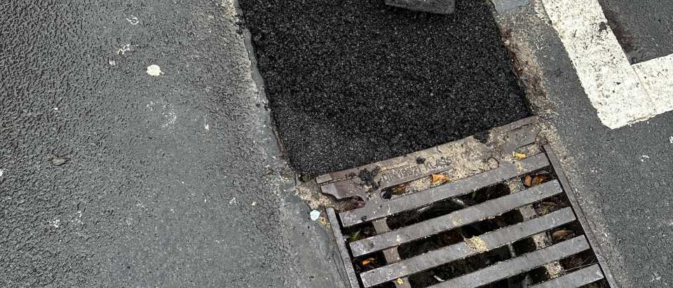 Mitarbeiter glättet frisch eingebauten Asphalt direkt neben einem Straßenablauf mit Gitterrost
