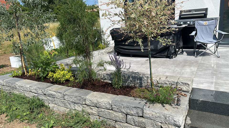 Niedrige Natursteinmauer mit bepflanztem Beet vor einer modernen Terrasse mit großformatigen Platten