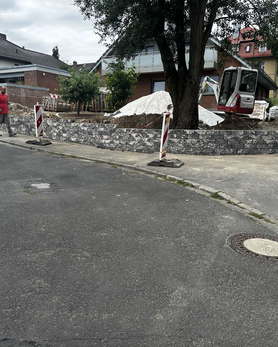 Neue Naturstein-Stützmauer entlang eines Grundstücks mit Minibagger und zwei Mitarbeitern auf der Baustelle