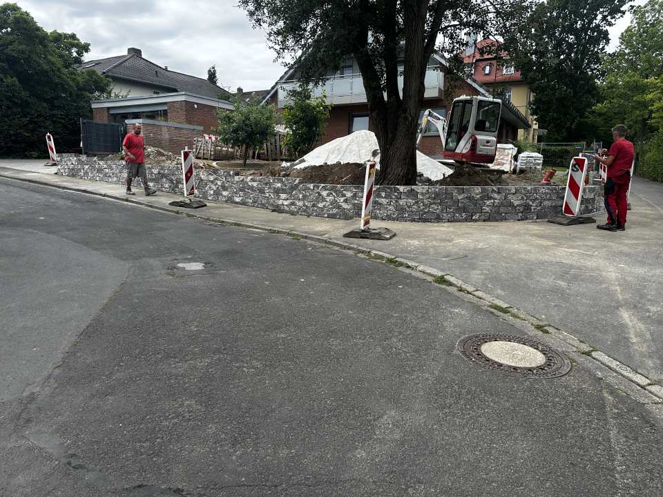 Neue Naturstein-Stützmauer entlang eines Grundstücks mit Minibagger und zwei Mitarbeitern auf der Baustelle