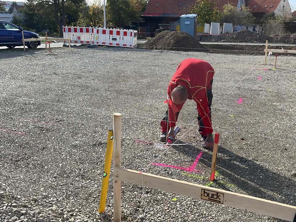 Mitarbeiter misst und markiert eine großflächig vorbereitete Schotterfläche mit Schnurgerüst und Holzpflöcken